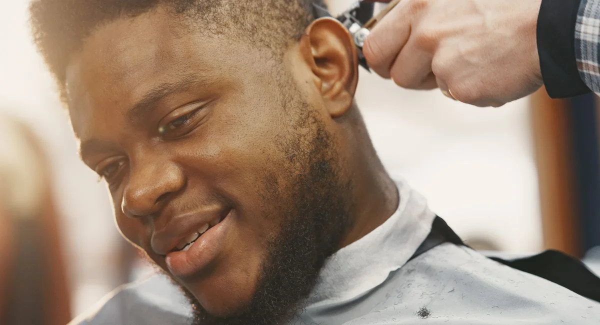 A close-up of a smiling black man getting a haircut at the barbershop indoors.