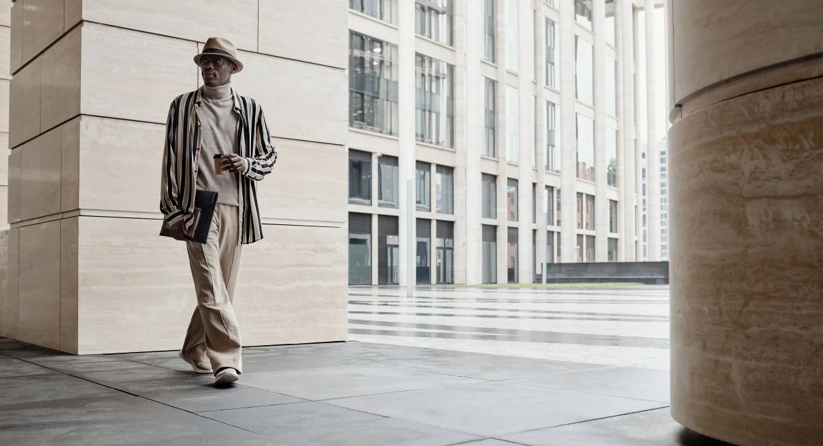 Fashionable man wearing hat and striped jacket walking in a modern cityscape.