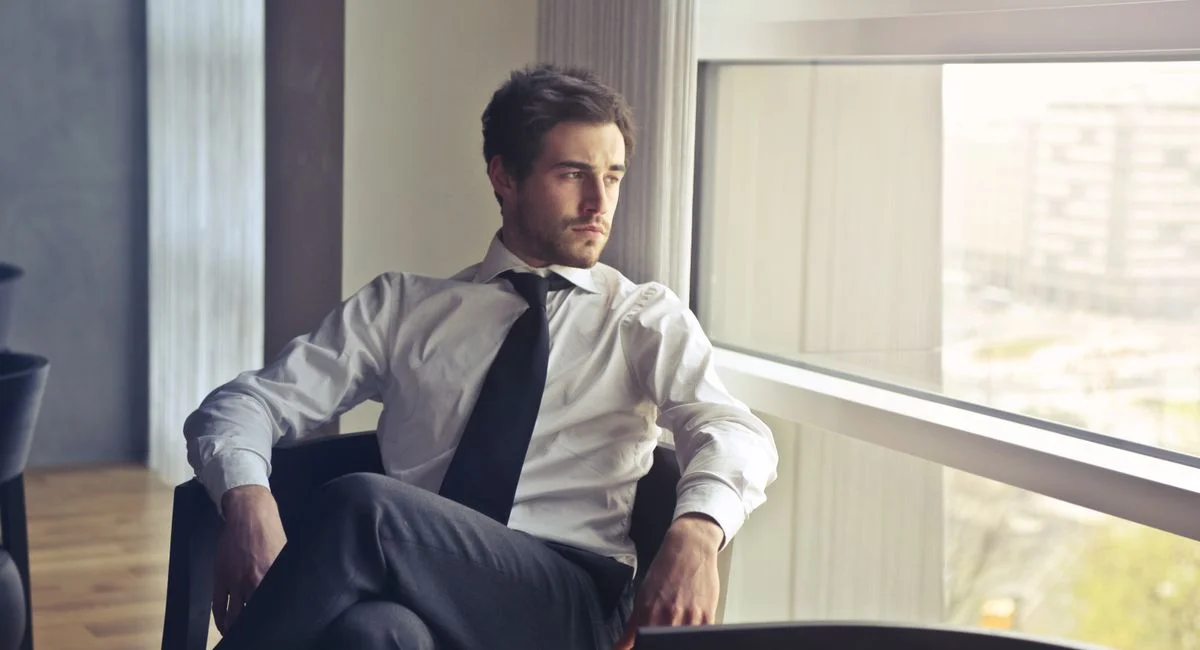 Young businessman in formal attire sitting and looking out of modern office window, contemplating.