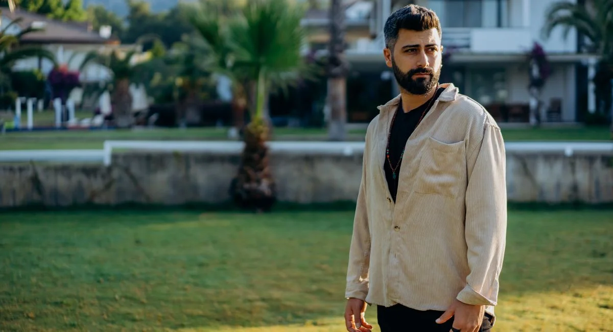 Stylish bearded man in casual attire standing outdoors with a scenic background.
