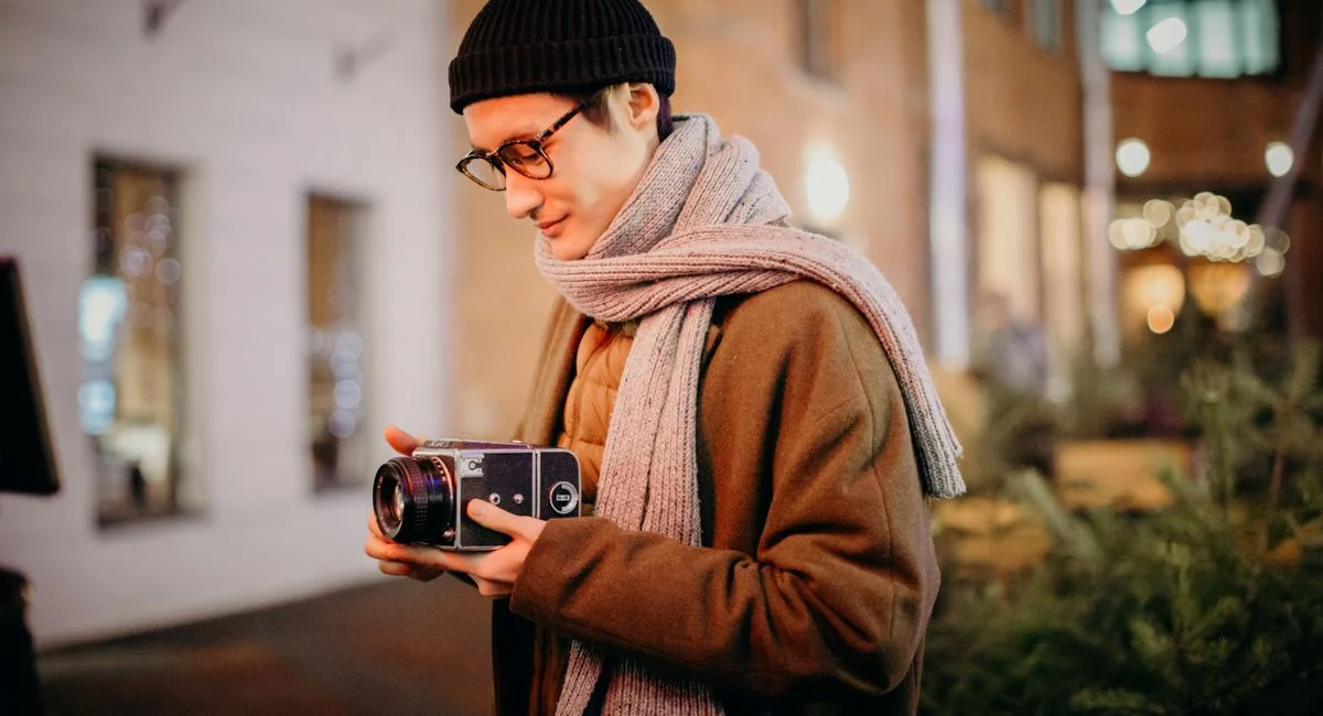 A young man in winter attire holding a vintage camera outdoors during a city evening.