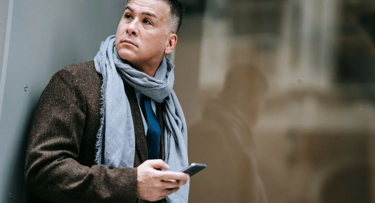 Man in formal outerwear with a scarf, holding a smartphone, outdoors with a reflective glass background.