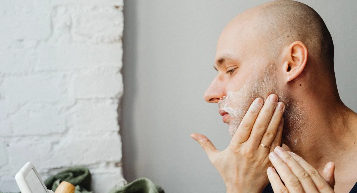 A bald man applies shaving cream to his face, focusing on personal grooming indoors.
