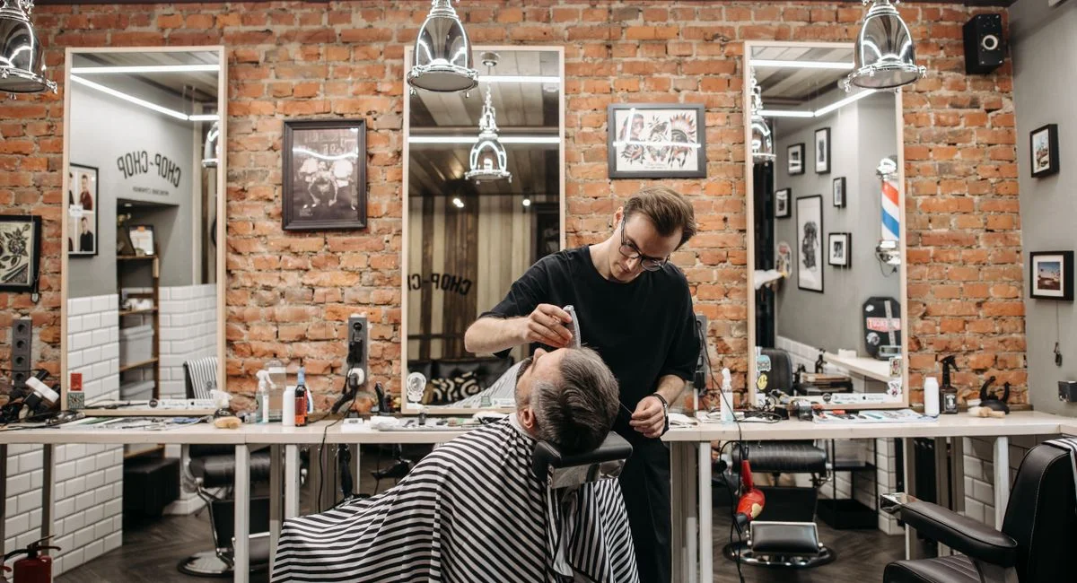 Barber working on a client's haircut in a stylish barbershop with brick walls and mirrors.