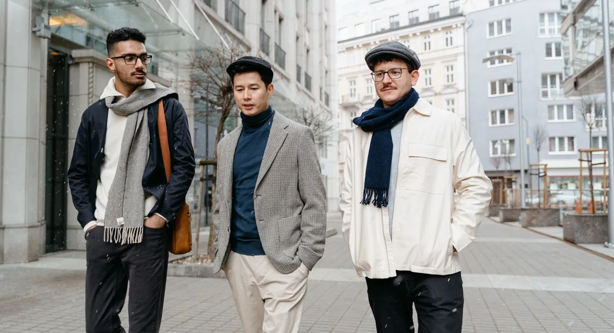 Three fashionable men stroll along a city street, showcasing modern winter style with scarves and flat caps.