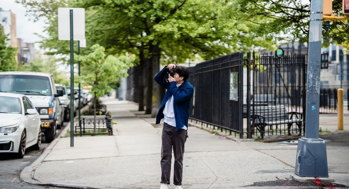 A man takes a photo on a city sidewalk surrounded by greenery and urban elements.