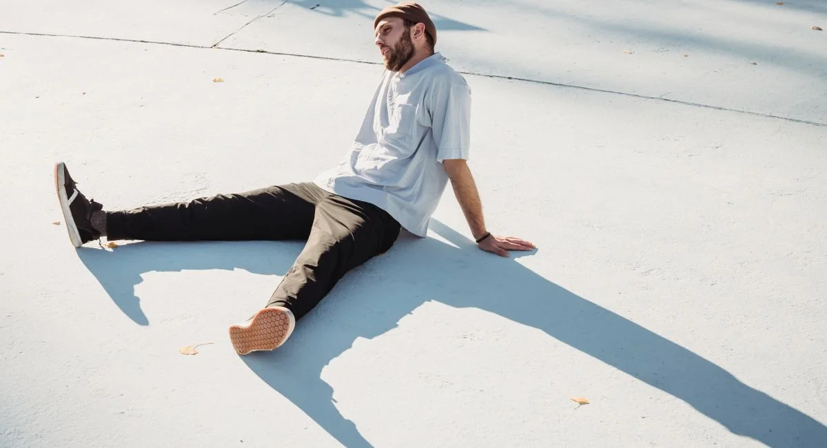 Young contemplative unshaven male in casual wear sitting on pavement with shadows and looking away in sunlight