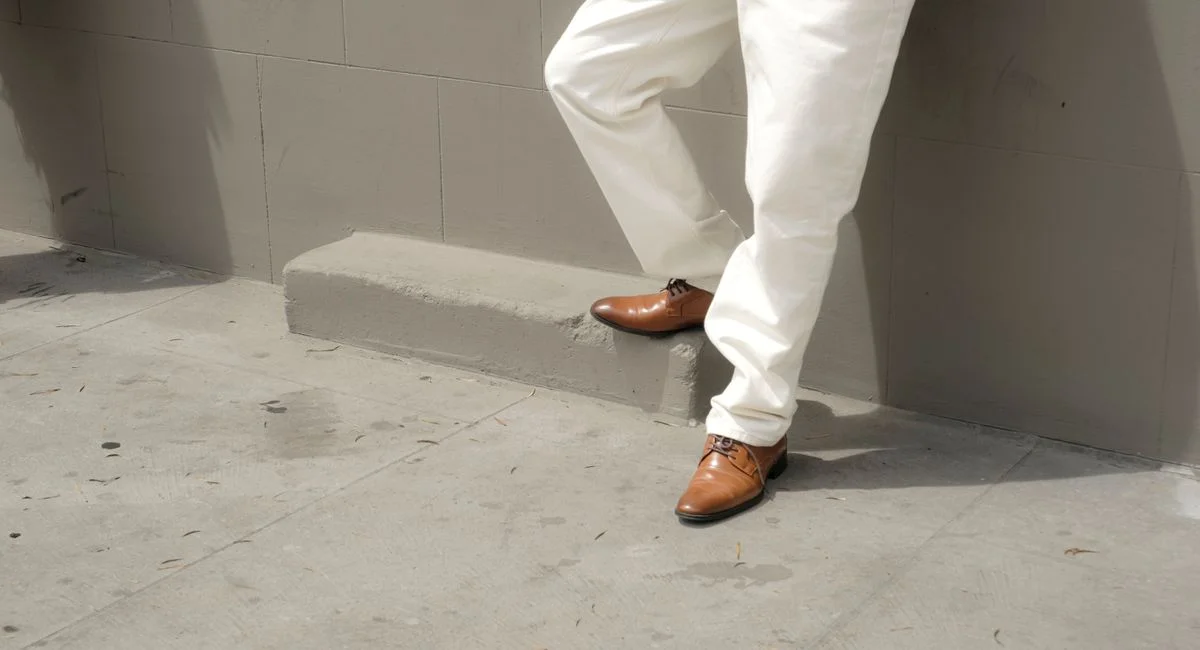 A person wearing classic brown Oxfords and white pants stands stylishly against a concrete urban wall.