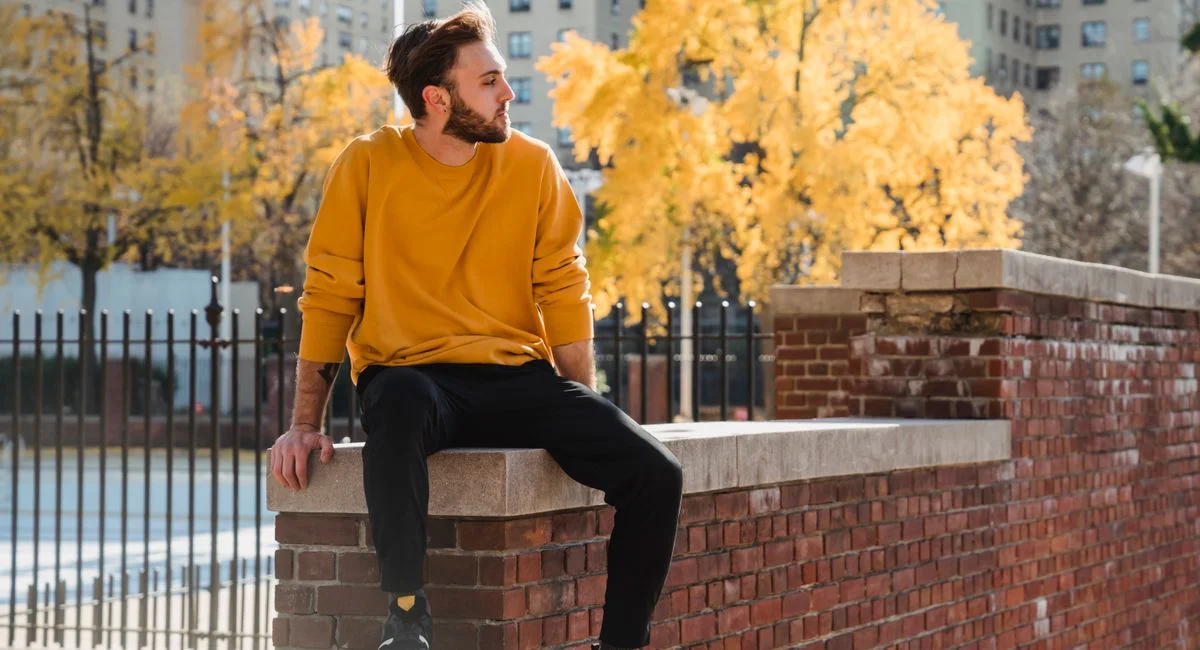 Side view of confident male sitting on brick fence and looking into distance on street against buildings with lush trees