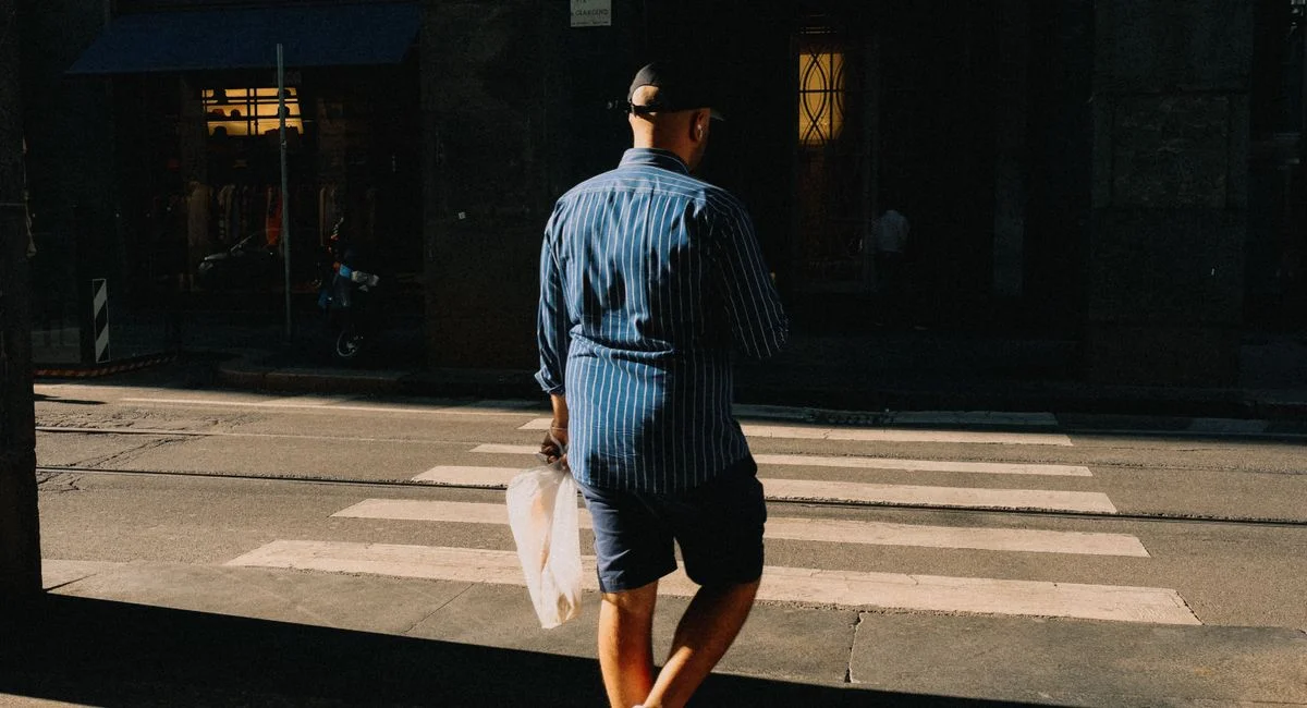 A man in striped attire crosses a sunlit street at a pedestrian crossing, holding a bag.