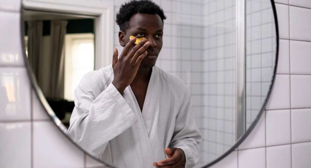 A man wearing a bathrobe applies an under-eye patch in front of a round mirror in a tiled bathroom.