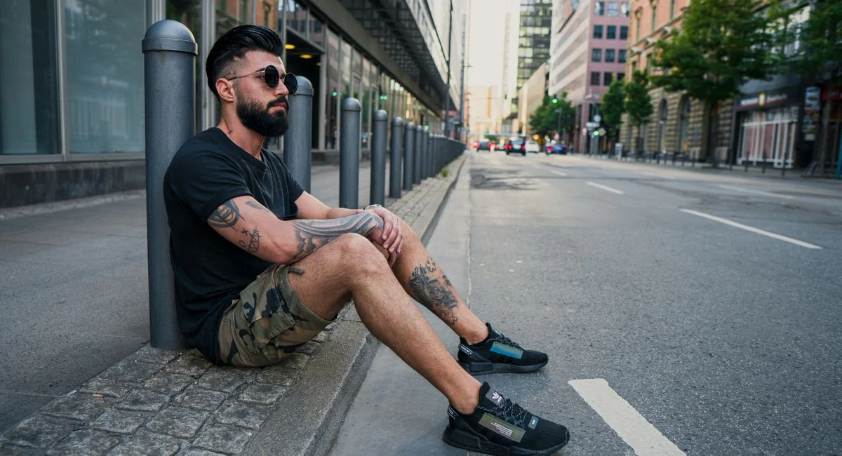 Side view of full body of bearded ethnic man in casual wear and sunglasses resting on sidewalk while sitting at pavement near